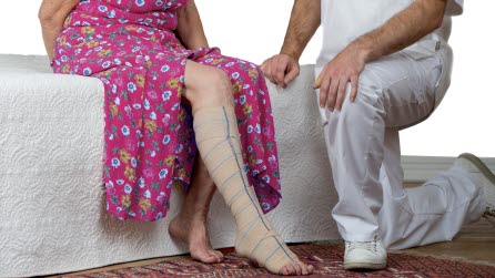 En kvinna i rosa klänning med ett bandage runt benet sitter på en säng. Bredvid henne sitter en läkare på huk.