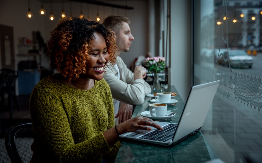 En kvinna sitter på ett café med en laptop framför sig. II bakgrunden syns en man med en kaffekopp framför sig.