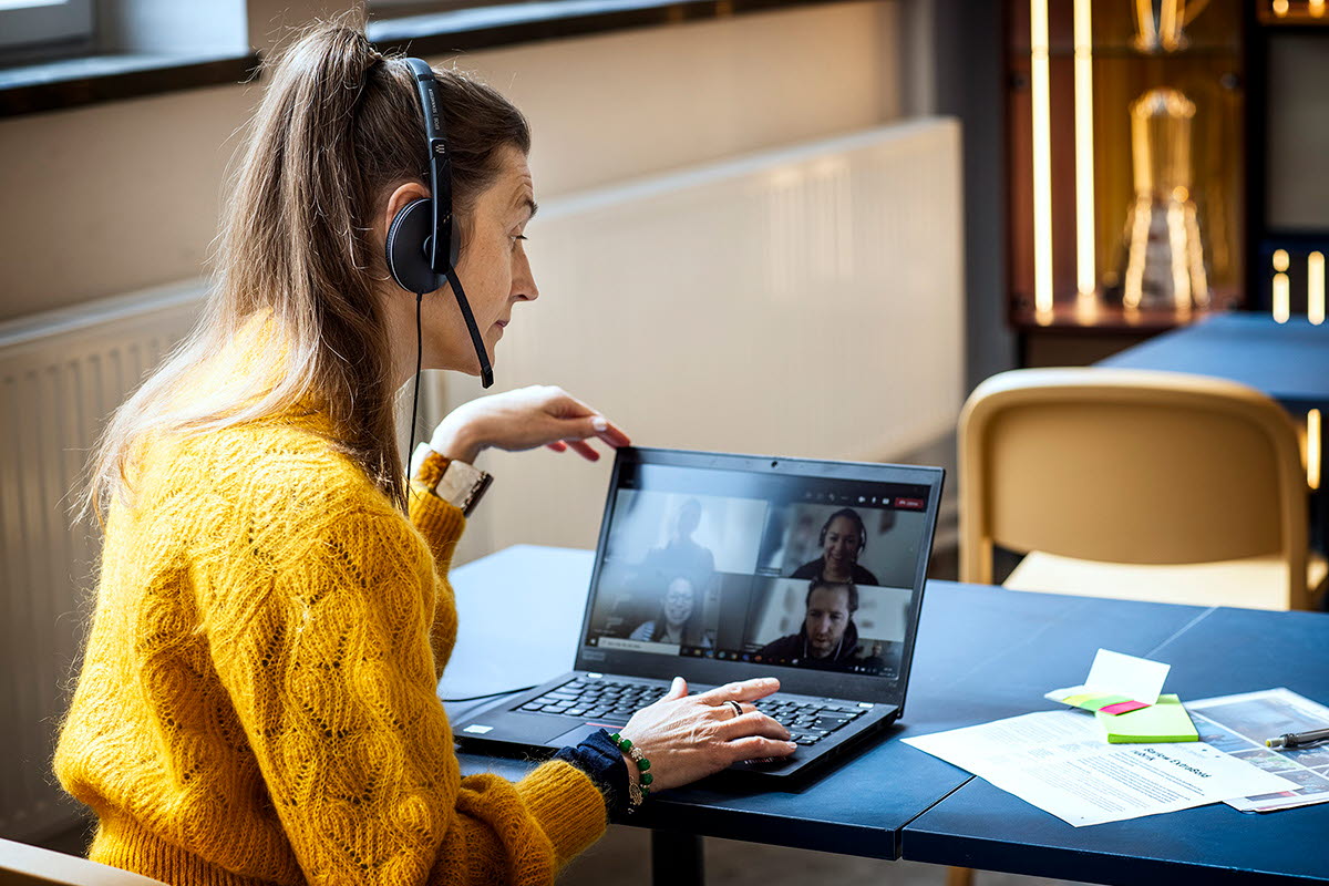 En kvinna i gul stickad tröja och hästsvans sitter med headseat vid en laptop och är inbegripen i ett samtal. 