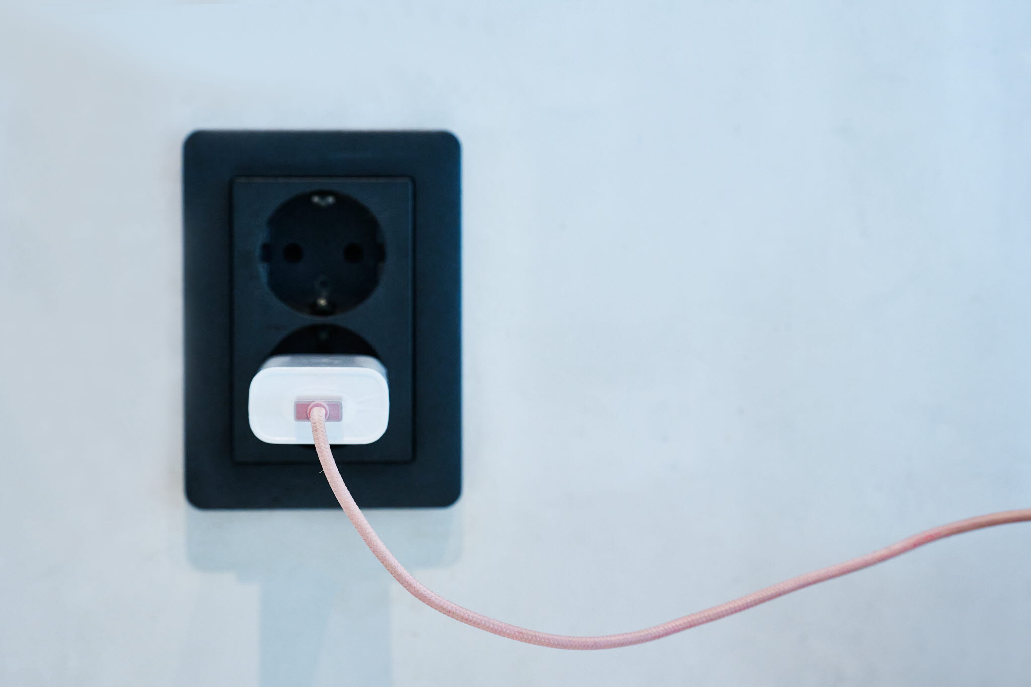 A white plug with a beige cord is plugged into a black wall socket.