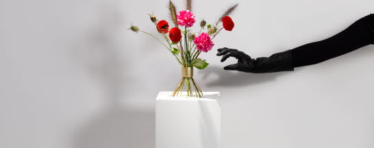 En blombukett står på en vit piedelstal. I bilden syns också en hand klädd i svart handske som försöker gripa tag i blombuketten.