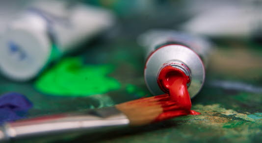 A paintbrush and paint tubes are seen lying on a table covered in paint.