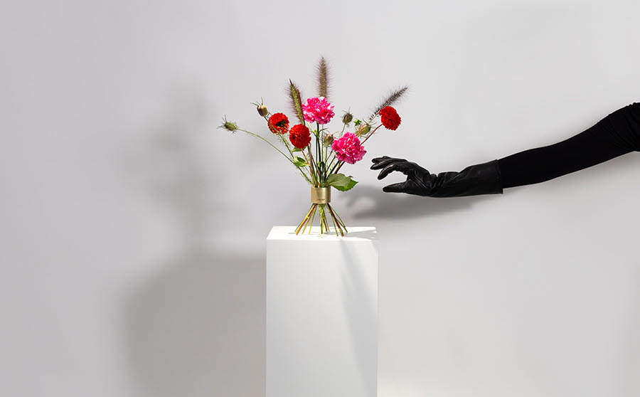 En blombukett står på en vit piedelstal. I bilden syns också en hand klädd i svart handske som försöker gripa tag i blombuketten. 